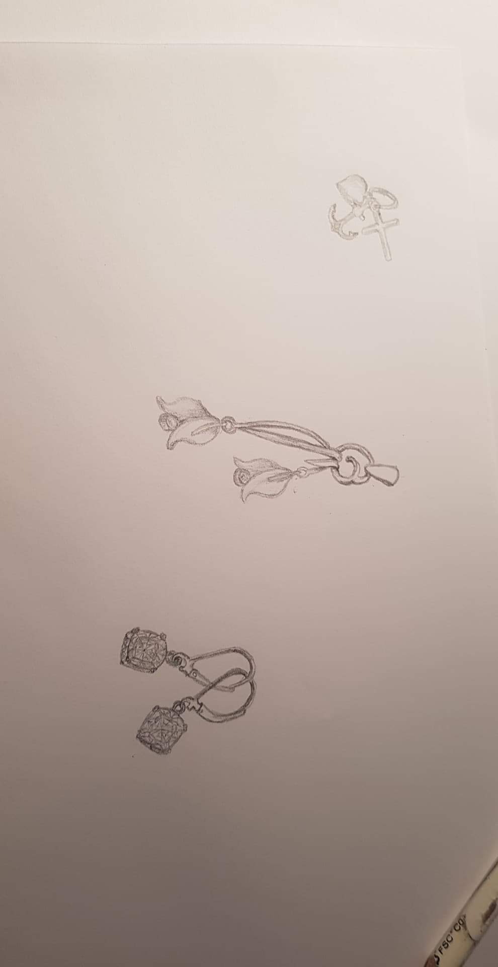 Szkic ołówkiem na papierze przedstawiający trzy projekty biżuterii: kolczyki z motywem kwiatowym, kolczyki z zawieszkami w kształcie kostek oraz zawieszkę z symbolem krzyża i kotwicy.