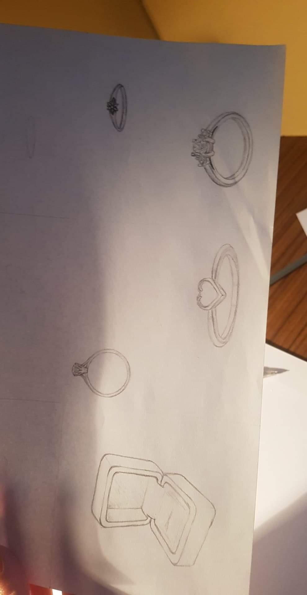 Szkic ołówkiem na papierze przedstawiający różne projekty pierścionków, w tym jeden z motywem serca i otwarte pudełko na pierścionek.