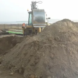 Żółta koparka w trakcie prac ziemnych przy budowie domu z drewnianą elewacją, widoczny wykop i hałda ziemi.