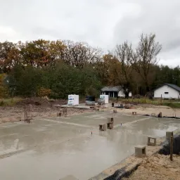 Wylany fundament pod budynek jednorodzinny, widoczne wystające pręty zbrojeniowe, bloczki betonowe, w tle drzewa i dwa budynki gospodarcze.