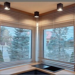 Verti- Room - Jasne żaluzje drewniane w kuchni narożnej, widok na ogród z sosnami, czarny okap, trzy lampy sufitowe.