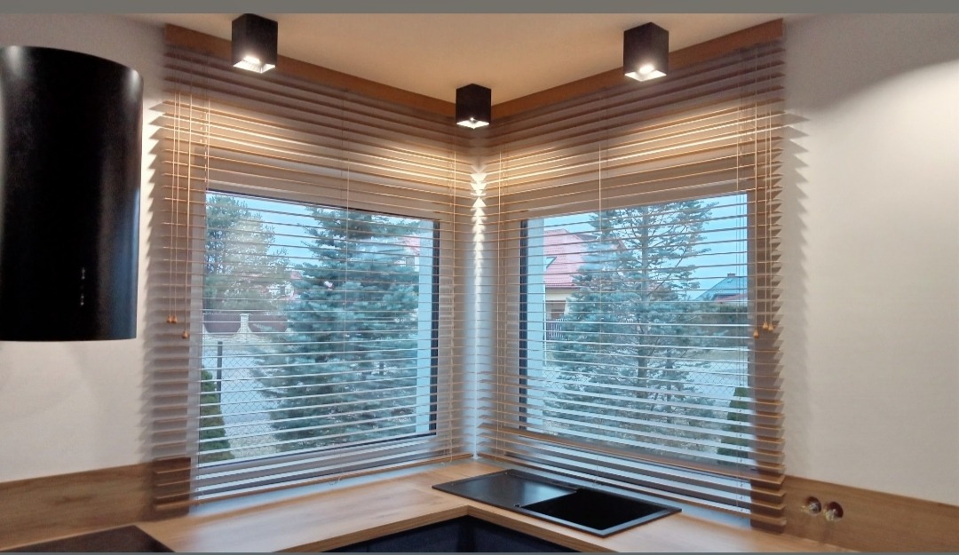 Jasne żaluzje drewniane w kuchni narożnej, widok na ogród z sosnami, czarny okap, trzy lampy sufitowe.