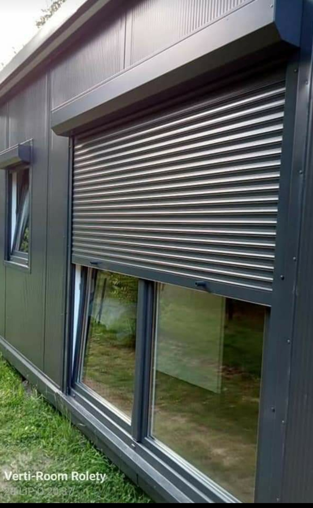 Częściowo opuszczona, szara roleta zewnętrzna na oknie budynku o metalowej elewacji, z widocznym fragmentem trawnika.