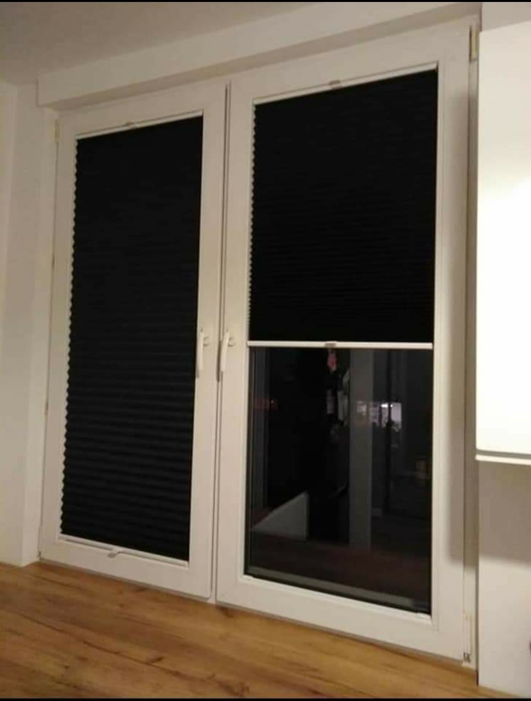 Czarne plisy okienne na białych, podwójnych drzwiach balkonowych z widocznym uchwytem i częściowo odsłoniętym oknem, na drewnianej podłodze.