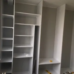 Wnętrze szafy wnękowej z białymi półkami, widoczne poziomice i narzędzia na półkach, surowy stan wykończenia.