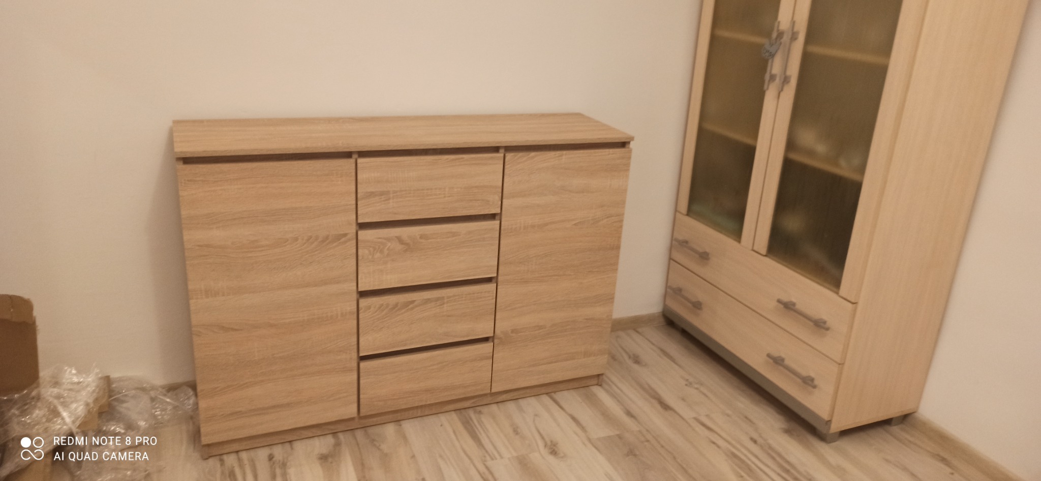 Komoda z trzema szufladami i szafka z przeszkleniami w odcieniach jasnego drewna, częściowo rozpakowane z folii i kartonu, ustawione przy ścianie.