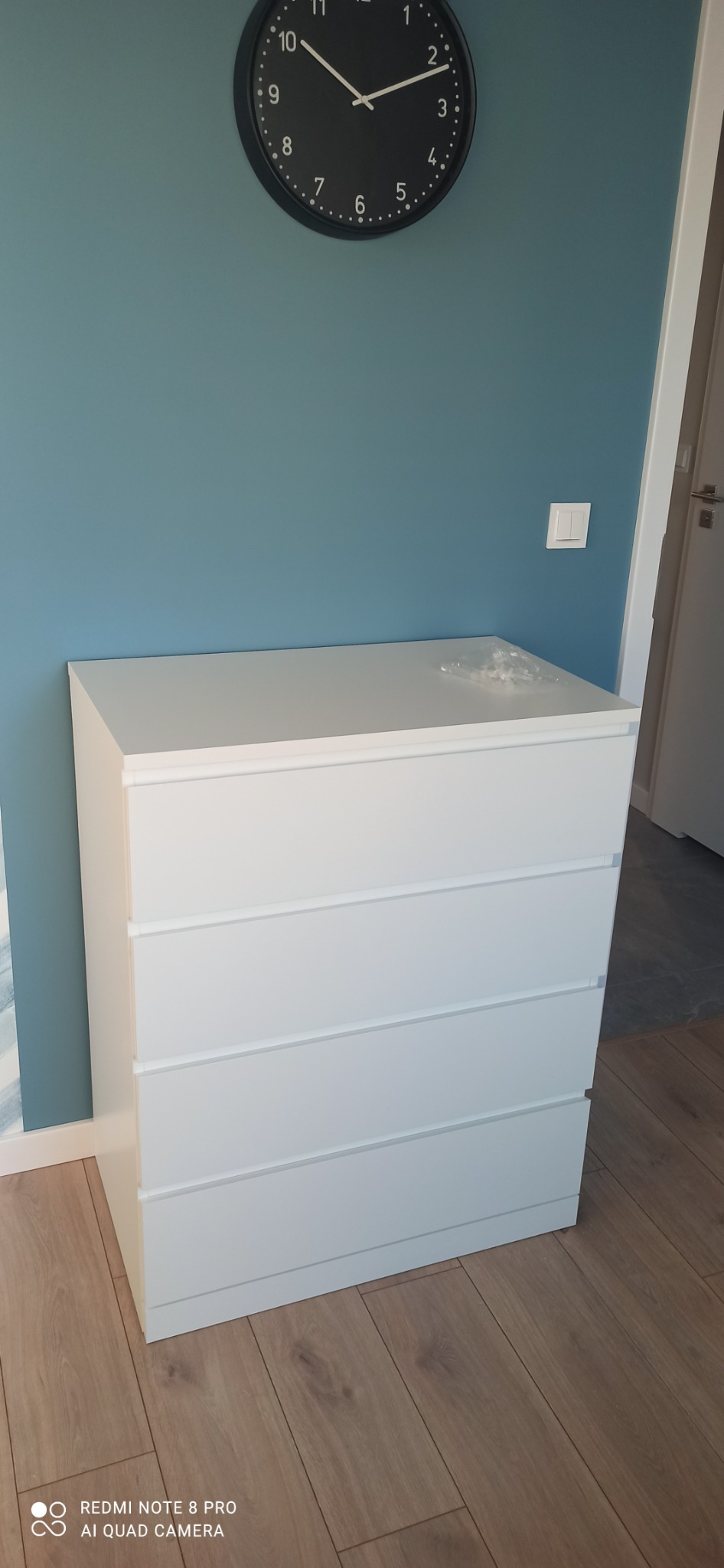 Biała komoda z czterema szufladami stoi na drewnianej podłodze przy błękitnej ścianie, nad nią wisi czarny zegar.