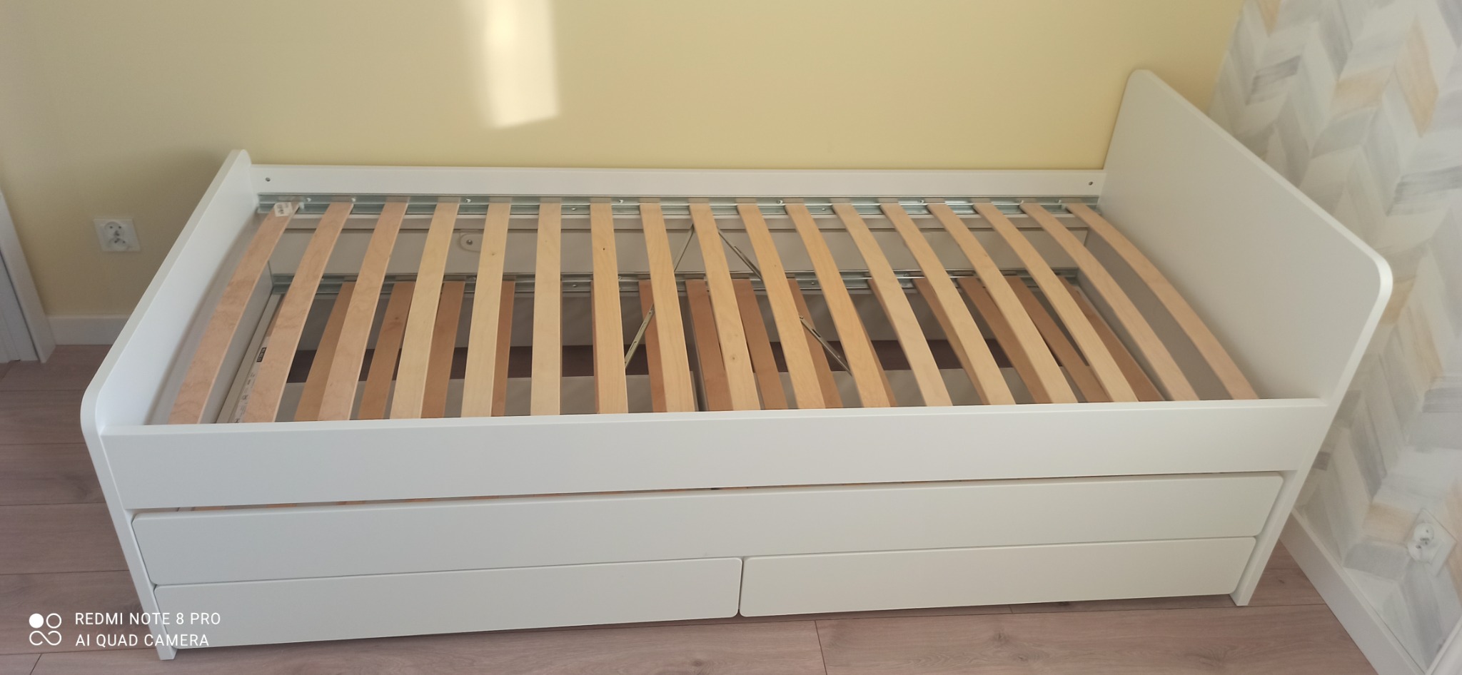 Biała rama łóżka z drewnianymi listwami i szufladami, widok z góry, z żółtą ścianą w tle i wzorzystą ścianą po prawej stronie.