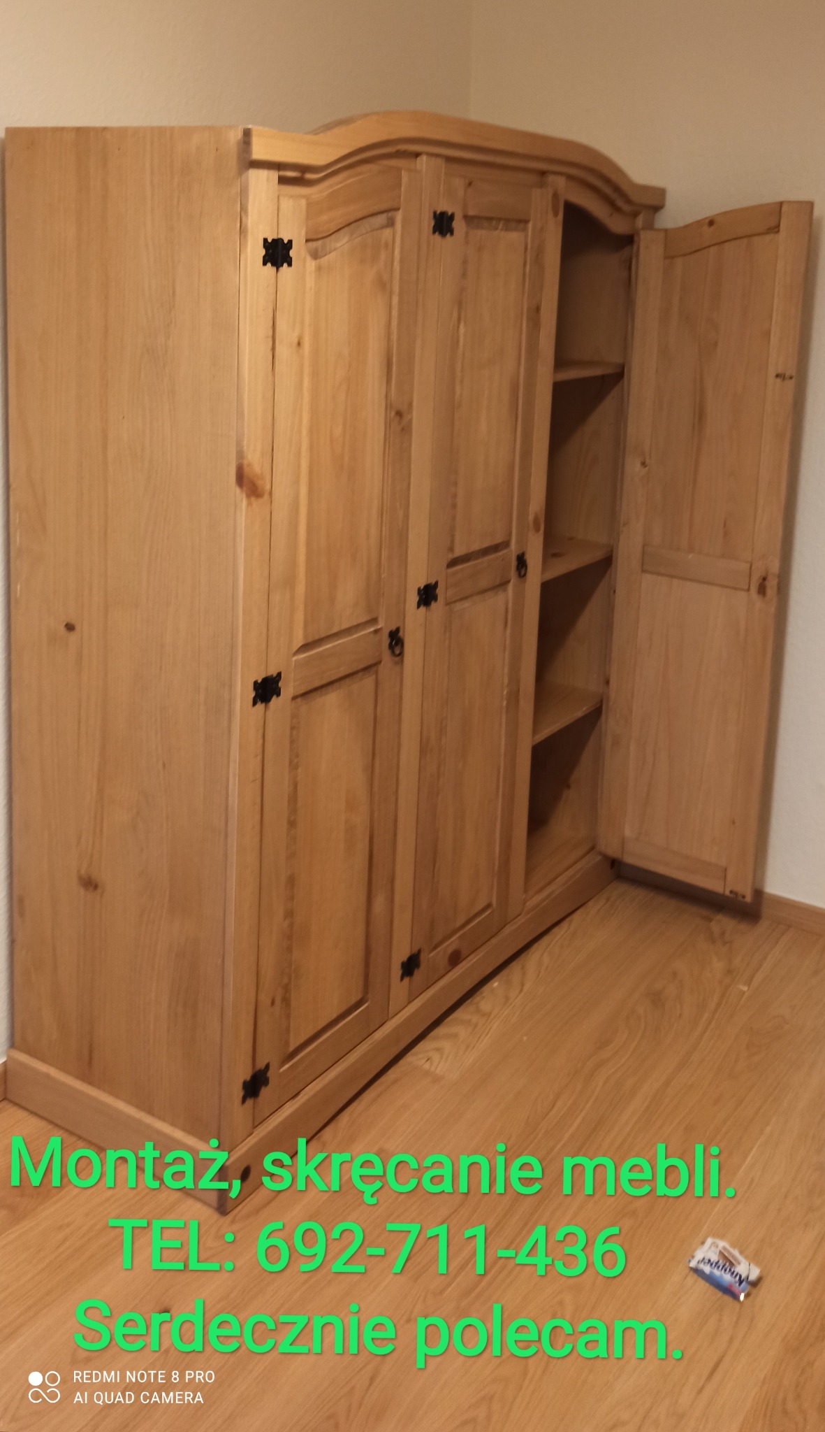 Drewniana szafa z trzema drzwiami, jedne drzwi otwarte, ukazujące półki wewnątrz, czarne okucia, widoczne napisy reklamowe na podłodze.