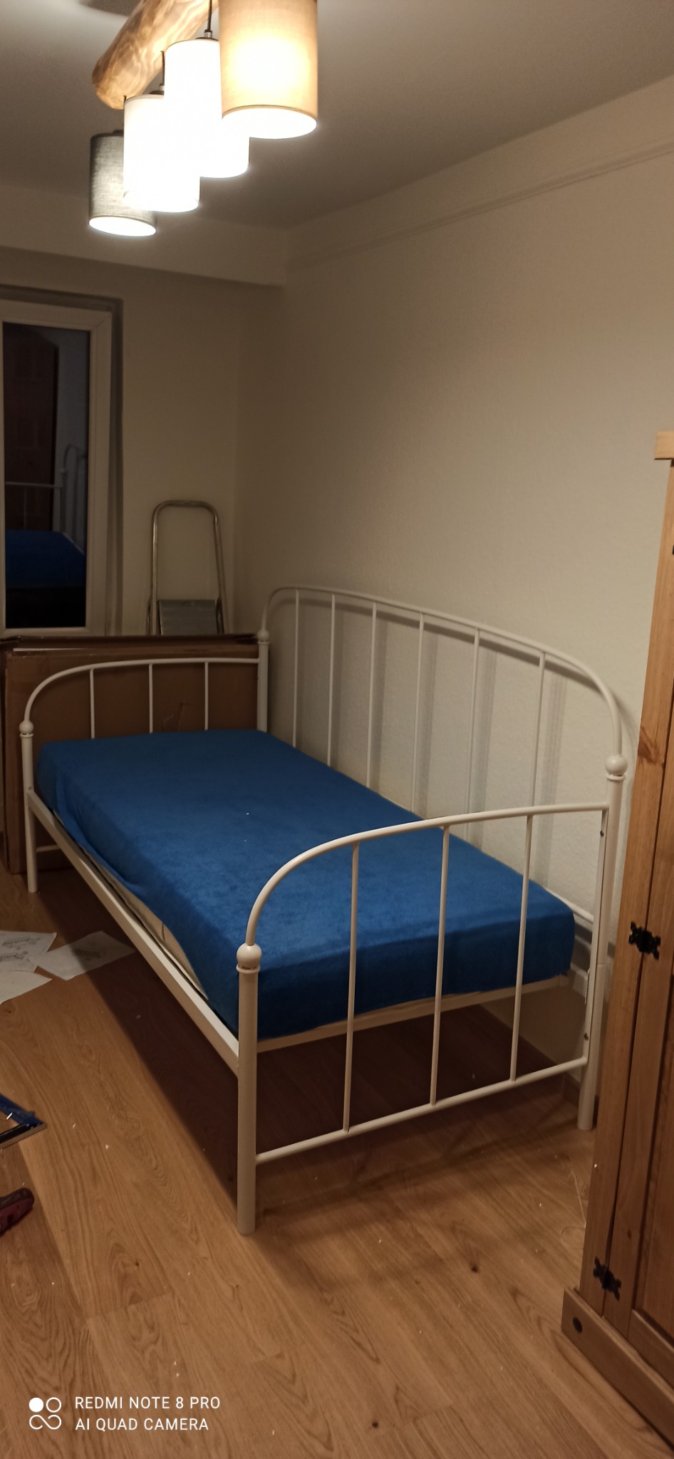 Biała metalowa kanapa z niebieskim materacem w pokoju, widoczna szafka i drabina w tle, oświetlenie sufitowe.