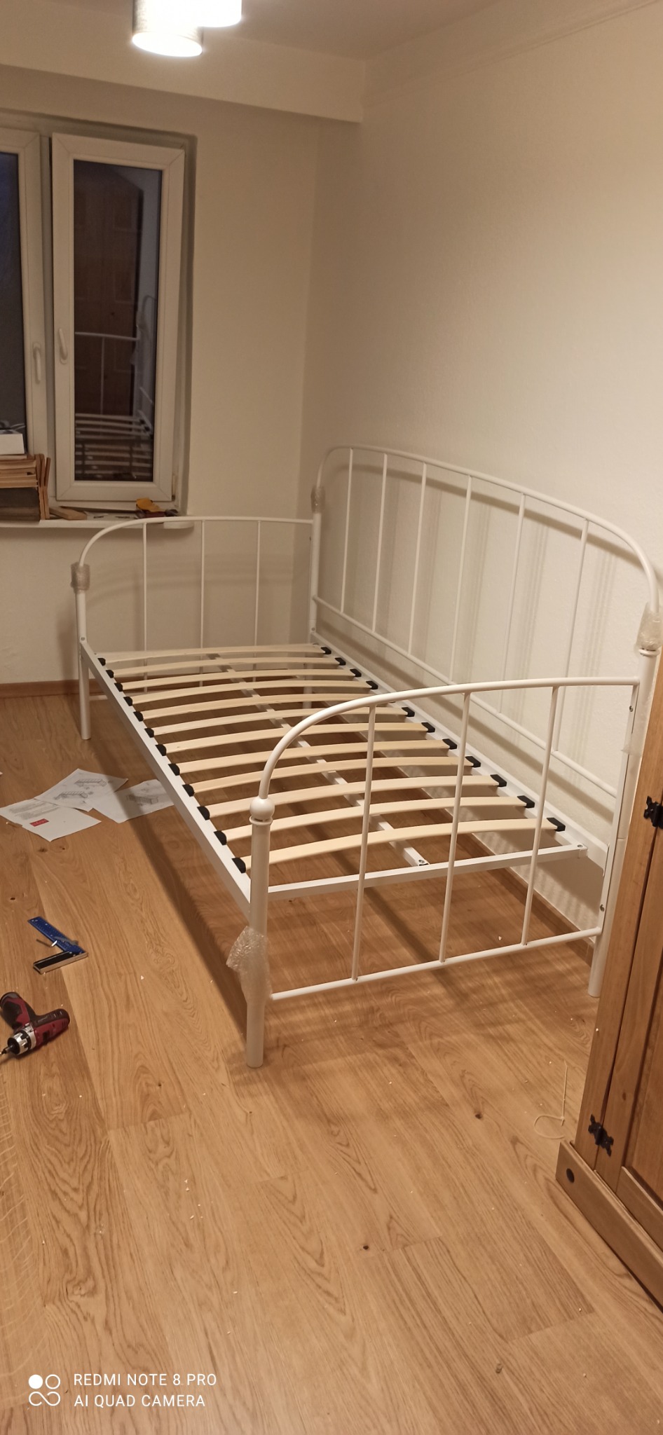 Biała metalowa rama łóżka z drewnianym stelażem, w trakcie montażu w pokoju z drewnianą podłogą, widoczne narzędzia i instrukcja.