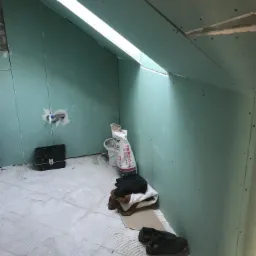 Poddasze w trakcie wykańczania płytami kartonowo-gipsowymi, widoczne łączenia płyt na skosach i resztki materiałów budowlanych na podłodze.