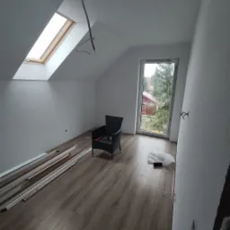 Poddasze po remoncie z oknem dachowym, drewnianą podłogą, listwami przypodłogowymi w rogu, czarnym fotelem i widokiem na ogród z okna.