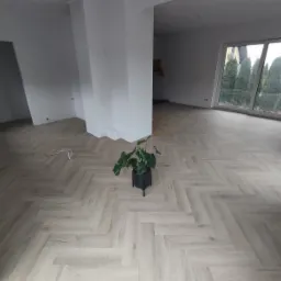 Nowoczesny salon z jasną podłogą ułożoną we wzór jodełki francuskiej, świeżo pomalowane ściany na biało, z widocznym fragmentem kolumny i dużym oknem balkonowym z widokiem na drzewa, doniczka...