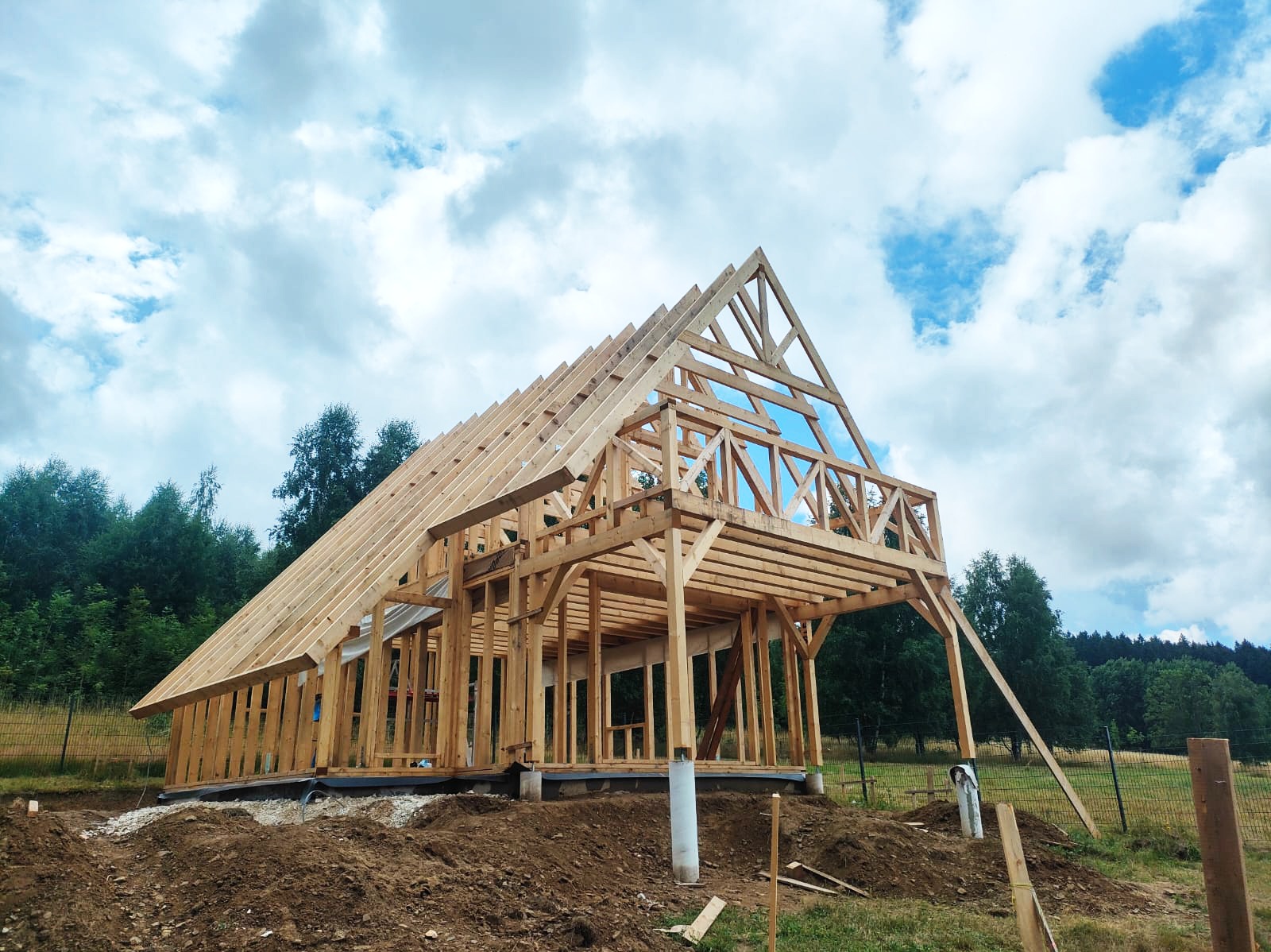 Surowy drewniany szkielet domu w trakcie budowy, widok z zewnątrz, z widoczną konstrukcją dachu i ścian, na tle zachmurzonego nieba i zieleni drzew.