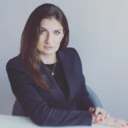 Portret kobiety w ciemnej marynarce, z długimi brązowymi włosami i niebieskimi oczami, siedzącej przy biurku z rękami złożonymi przed sobą, patrzącej w obiektyw.