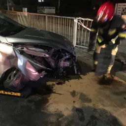 Uszkodzony srebrny samochód na żółtej platformie, strażak w czerwonym hełmie w pobliżu, nocne oświetlenie uliczne.