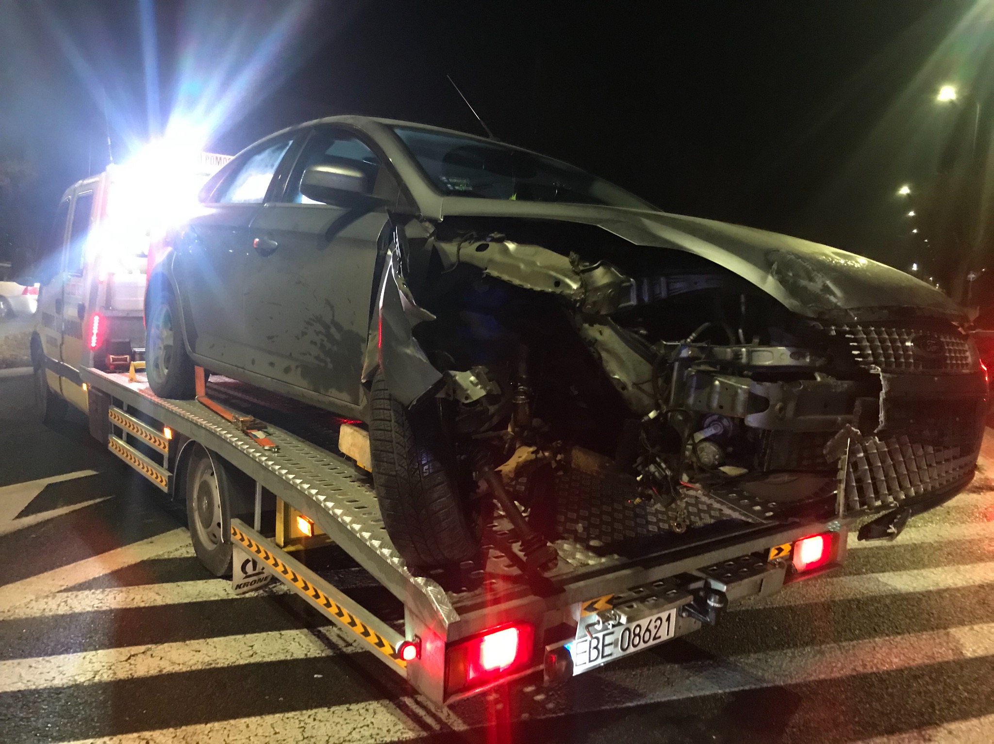 Uszkodzony szary samochód osobowy na lawecie w nocy, widoczne uszkodzenia przodu pojazdu i tablica rejestracyjna BE 08621.