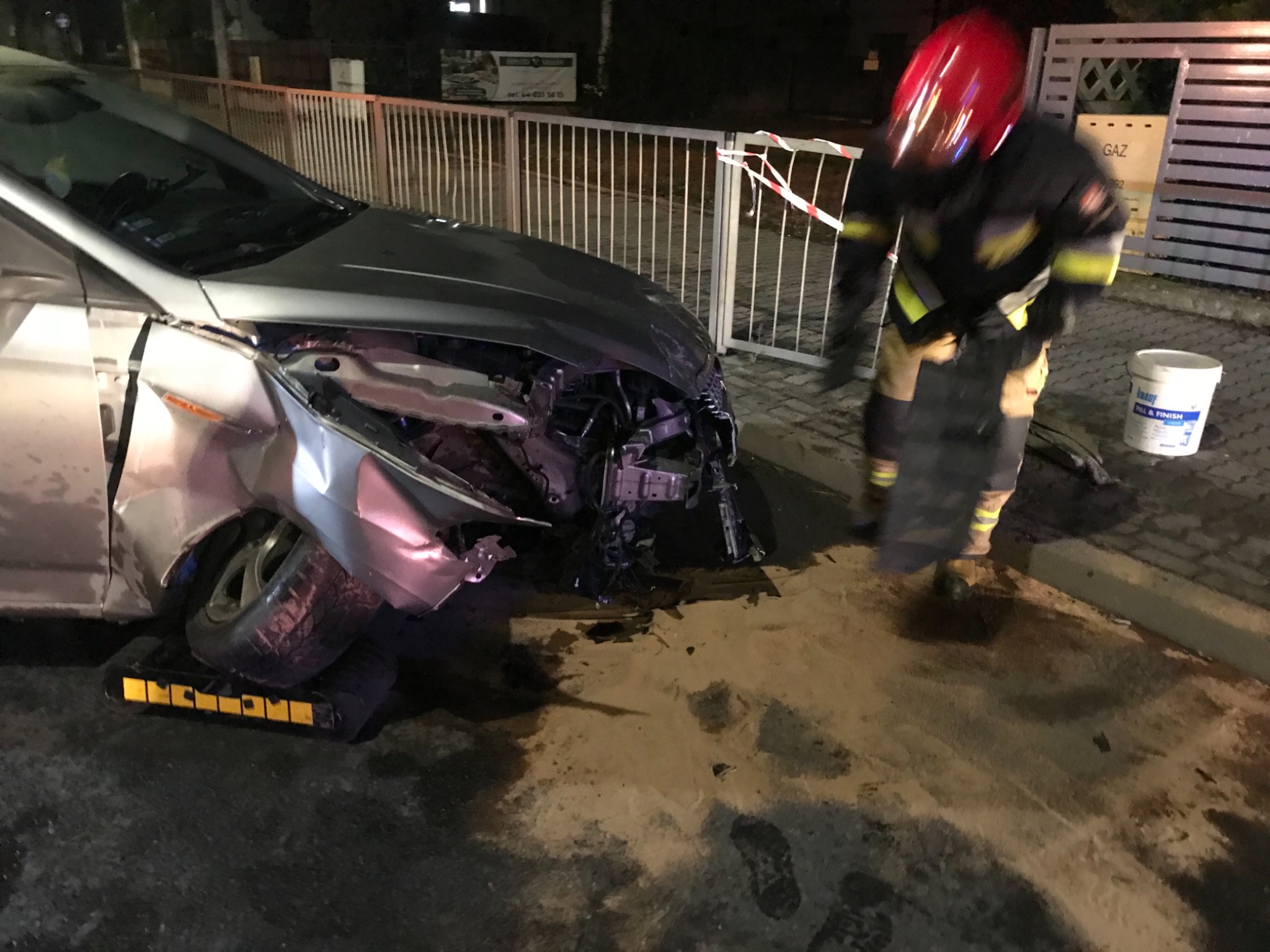 Uszkodzony srebrny samochód na żółtej platformie, strażak w czerwonym hełmie w pobliżu, nocne oświetlenie uliczne.