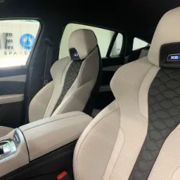 Wnętrze luksusowego samochodu BMW X6 z jasną skórzaną tapicerką, widoczne fotele z perforowanej skóry w kolorze białym i czarnym o strukturze plastra miodu, zagłówki z podświetlanym logo X6, widok...