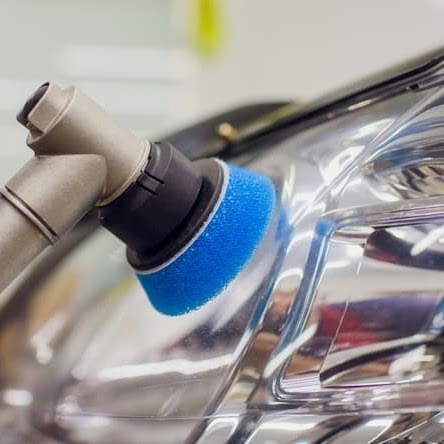 Detail of blue polishing pad attached to a tool, being used on a car headlight, reflecting light.
