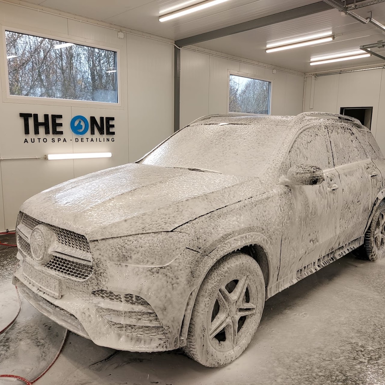 Mercedes-Benz GLE pokryty grubą warstwą aktywnej piany w jasnym pomieszczeniu myjni samochodowej, widoczne logo 'The ONE Auto Spa Detailing' na ścianie.