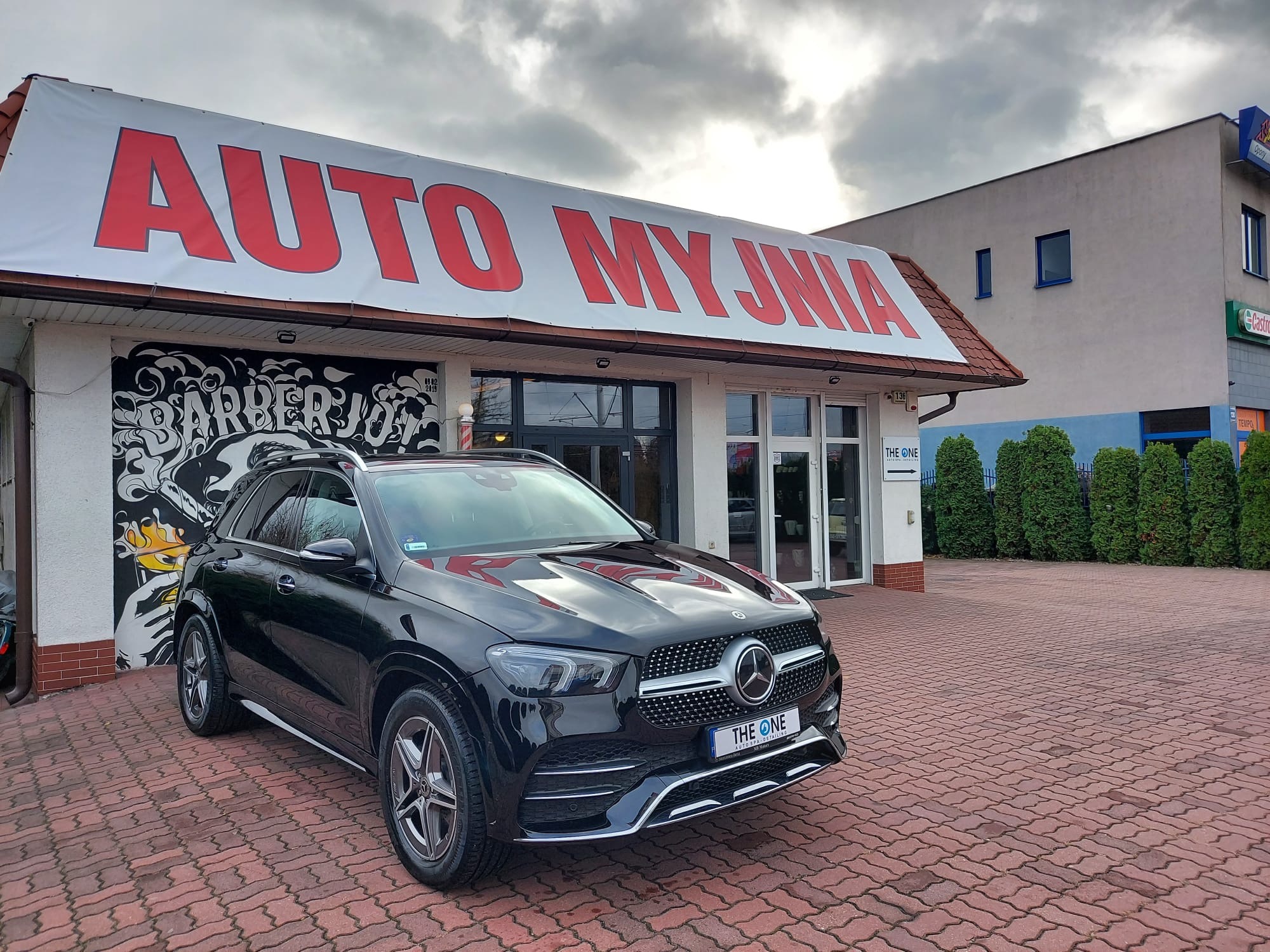 Czarny Mercedes GLE zaparkowany przed budynkiem myjni samochodowej z dużym banerem 'AUTO MYJNIA' na dachu i graffiti 'BARBERJO' na ścianie obok wejścia.