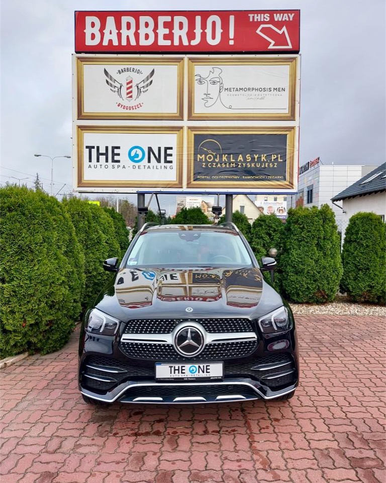 Czarny Mercedes GLE po detailingu, zaparkowany na kostce brukowej przed budynkiem z reklamami salonów barberskich i auto spa, widoczne odbicie billboardu w lakierze maski.