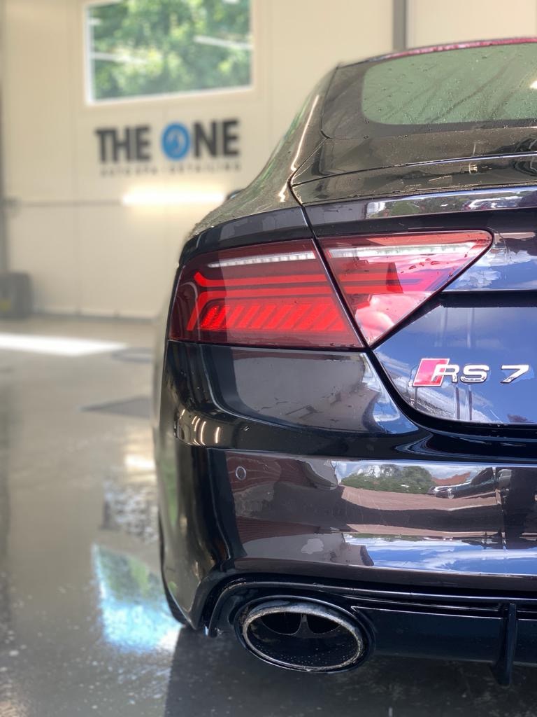 Tył czarnego samochodu Audi RS7 z kroplami wody, widoczny pojedynczy wylot wydechu, logo RS7 i tylne światła, w tle okno i napis 'THE ONE'.