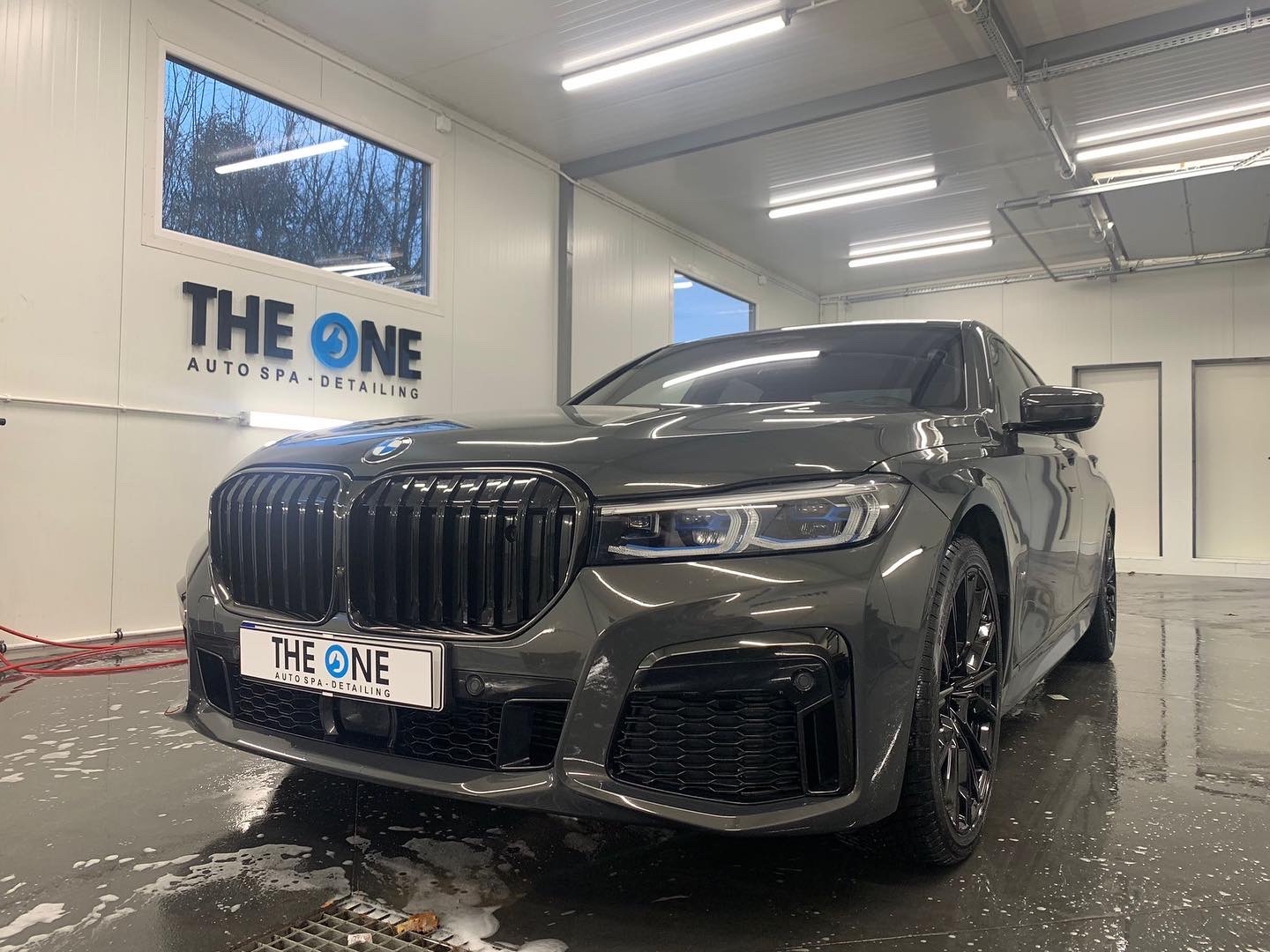 Ciemnoszary BMW serii 7 po myciu w myjni detailingowej, widoczny znak 'THE ONE AUTO SPA - DETAILING' na ścianie i tablicy rejestracyjnej, mokra posadzka z odpływem