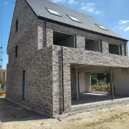 Dwukondygnacyjny dom w stanie surowym, z szarymi cegłami i ciemnoszarą dachówką, widoczne otwory okienne i fragment żurawia budowlanego po lewej stronie.