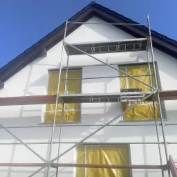 Biała elewacja domu jednorodzinnego z częściowo zakrytymi oknami folią ochronną i rozstawionym rusztowaniem, przygotowywana do prac wykończeniowych.