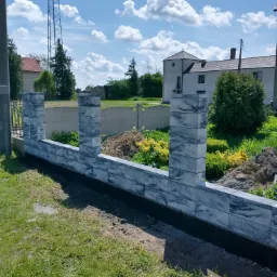 Wznoszony mur z szarych, blokowych elementów z widocznymi filarami, na tle zieleni i błękitnego nieba z chmurami.