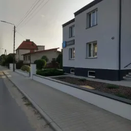 Odnowiony budynek dwukondygnacyjny z numerem 42, pomalowany na szaro z ciemnoszarym pasem u dołu, widoczny z chodnika wyłożonego szarą kostką brukową, z przodu rabata z krzewami, w tle inne budynki.