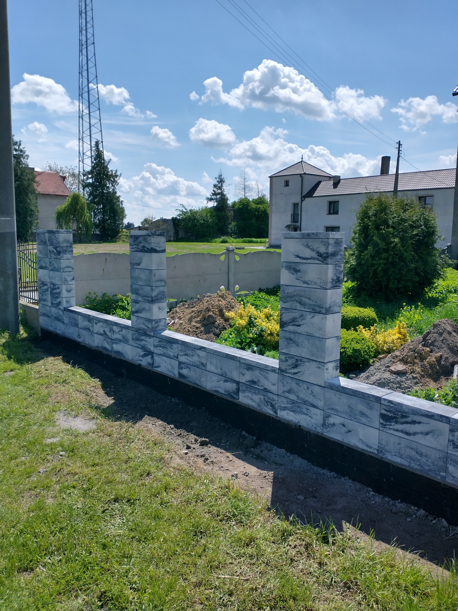 Wznoszony mur z szarych, blokowych elementów z widocznymi filarami, na tle zieleni i błękitnego nieba z chmurami.