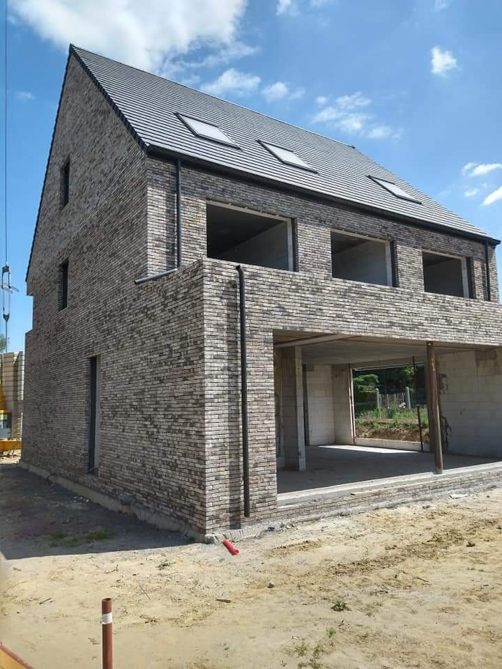 Dwukondygnacyjny dom w trakcie budowy z szarej cegły klinkierowej, widoczne otwory okienne bez ram, czarny dach z oknami połaciowymi, plac budowy z ziemią.