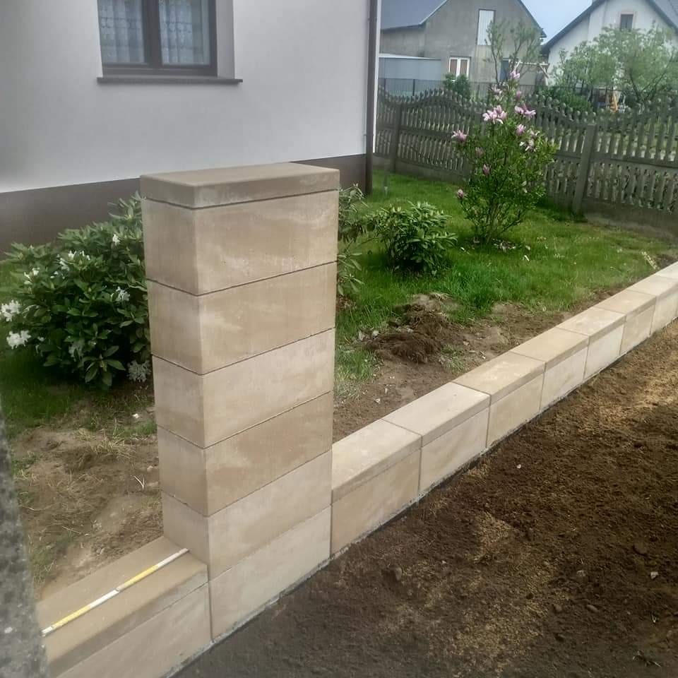 Wzniesiony z beżowych bloczków murek oporowy z filarem na tle trawnika z kwitnącymi krzewami i domem jednorodzinnym.