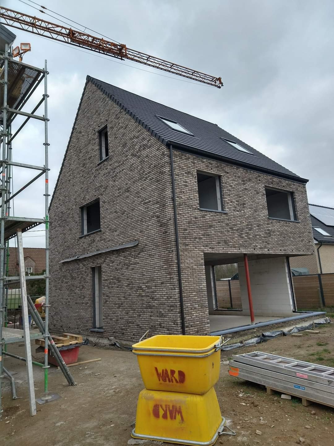 Surowy stan domu z szarej cegły z czarnym dachem i oknami bez szyb, widoczne rusztowania, żuraw budowlany i żółte wiadra z napisem 'WARD'.
