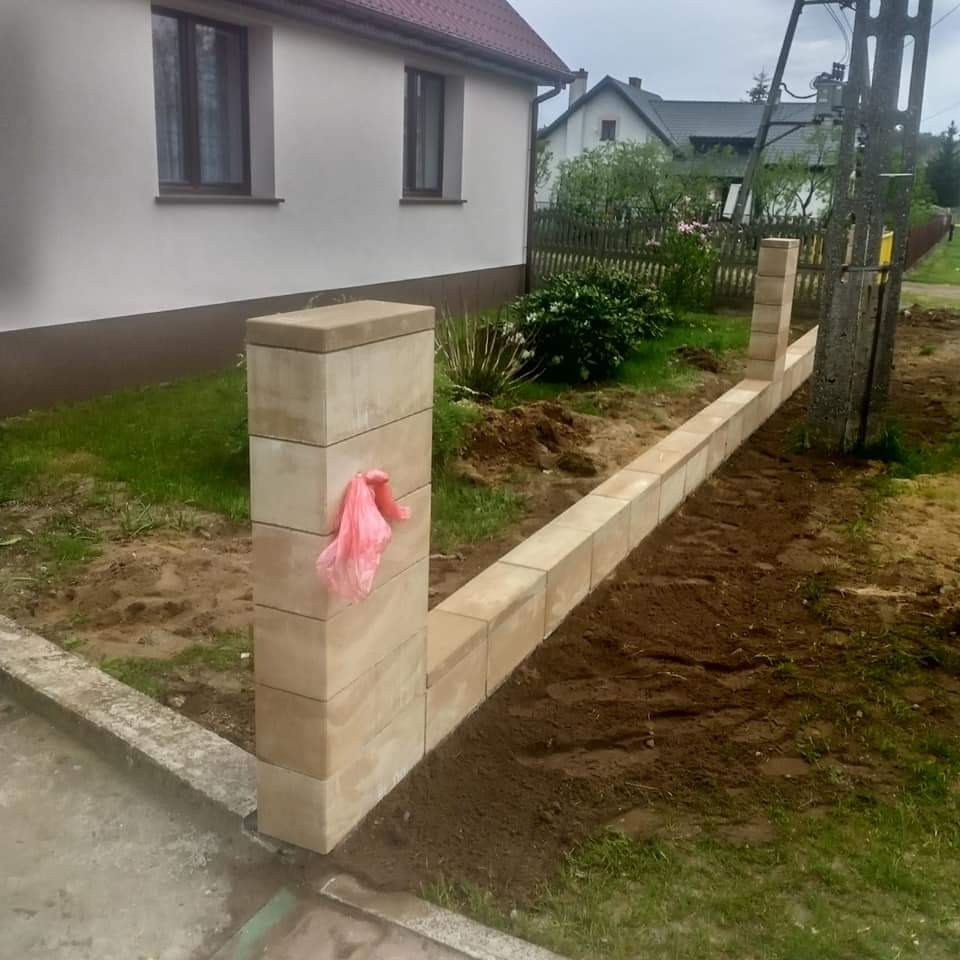 Wznoszona z beżowych bloczków ściana ogrodzenia na fundamencie, z widocznym fragmentem domu w tle i różową reklamówką przyczepioną do jednego z filarów.