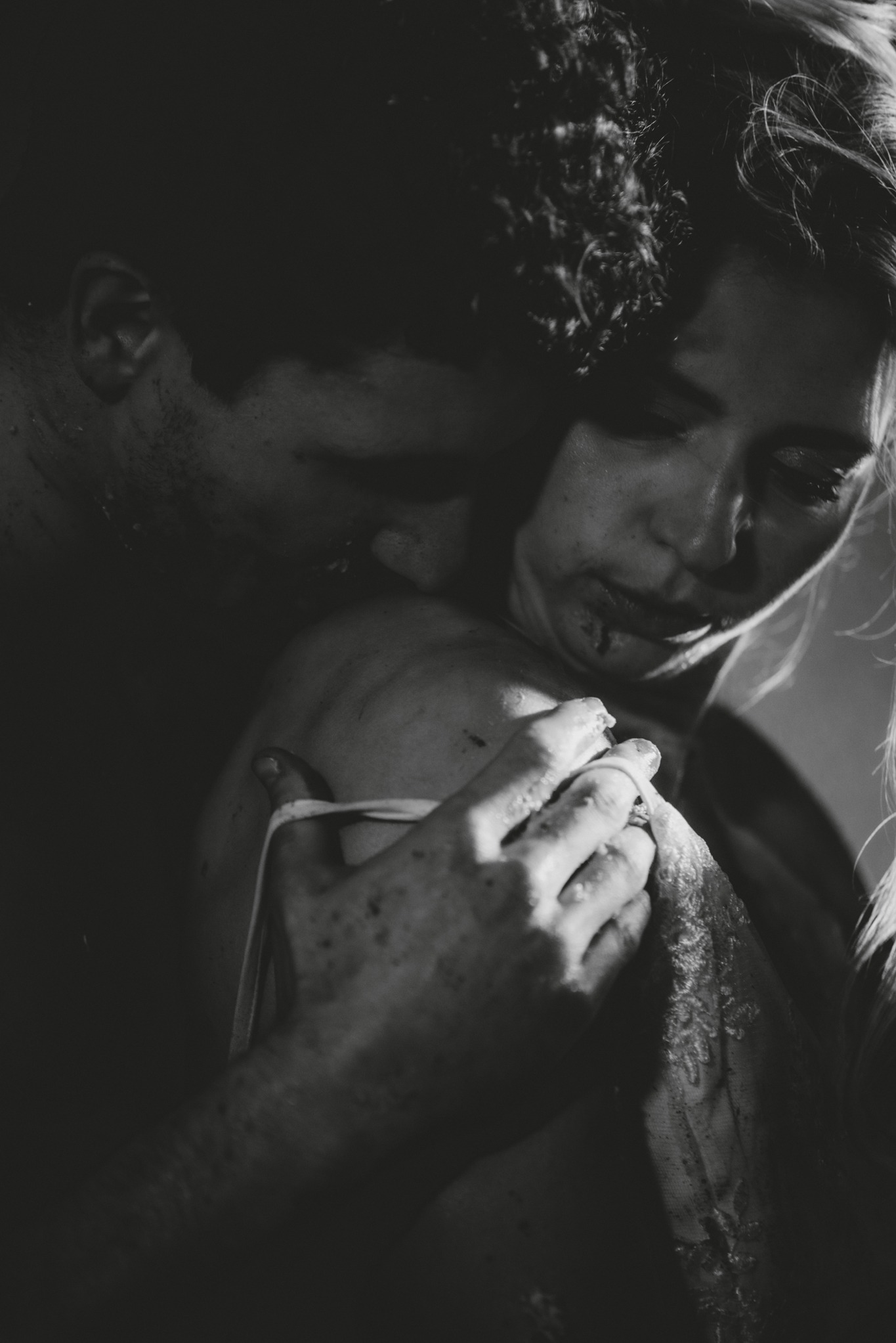 Czarno-białe, intymne ujęcie pary: mężczyzna obejmuje kobietę, delikatnie całując jej ramię, ona patrzy w dół z zamkniętymi oczami, widoczna faktura koronki na jej ubraniu i krople na twarzy.