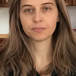 Portret kobiety z długimi, brązowymi włosami, ubranej w ciemny sweter z zamkiem, na tle drewnianych szafek kuchennych.