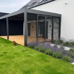 Nowoczesna pergola z drewna i metalu, z lamelami, przylegająca do białego domu z czarnymi akcentami w Lesznie. Na pierwszym planie trawnik z kwitnącą lawendą i pies.