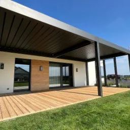 Nowoczesna pergola z drewnianym tarasem, zintegrowana z budynkiem, widok na zadbany trawnik i lawendę, błękitne niebo w tle.