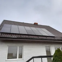 Panele słoneczne zamontowane na brązowym dachu dwuspadowym domu z białą elewacją sidingową, widoczne okna i fragment schodów wejściowych.