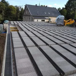 Budowa stropu Teriva: prefabrykowane belki i pustaki betonowe ułożone na ścianach fundamentowych, widoczne zbrojenie i częściowo deskowanie szalunkowe, w tle dom i maszyna budowlana.