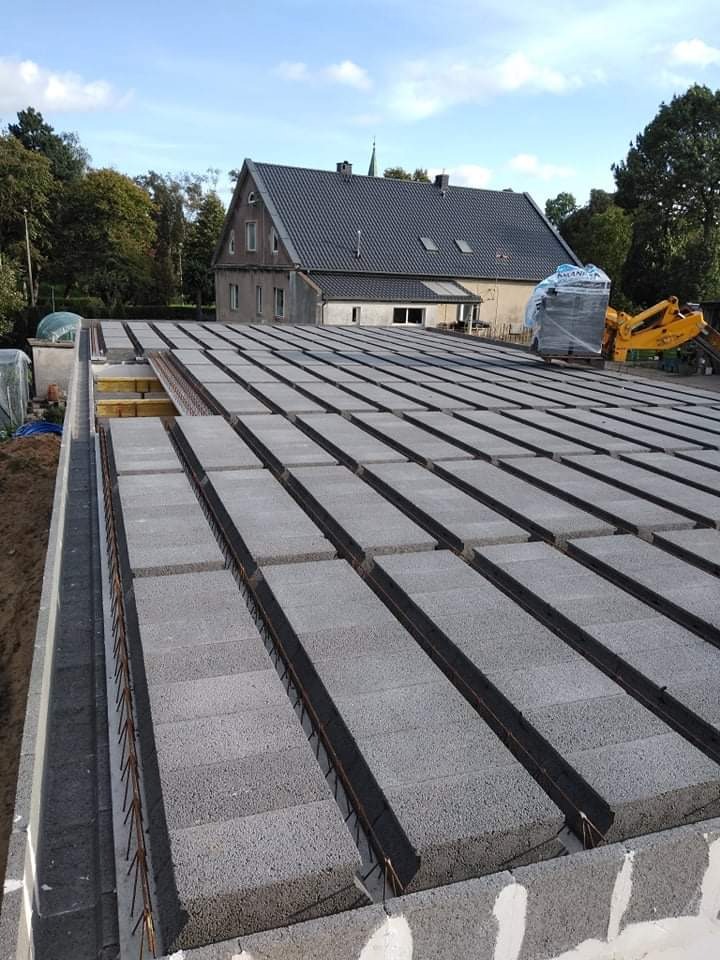 Budowa stropu Teriva: prefabrykowane belki i pustaki betonowe ułożone na ścianach fundamentowych, widoczne zbrojenie i częściowo deskowanie szalunkowe, w tle dom i maszyna budowlana.