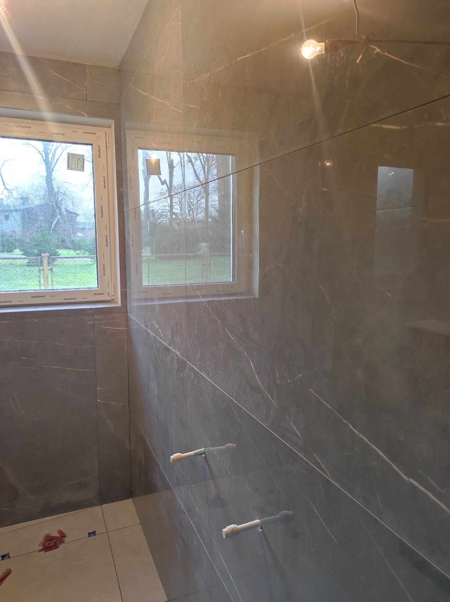 Ściana i podłoga w łazience wyłożone szarymi płytkami imitującymi marmur, widoczne okna z widokiem na zewnątrz i elementy mocujące płytki na ścianie, żarówka pod sufitem, podłoga częściowo wyłożona...
