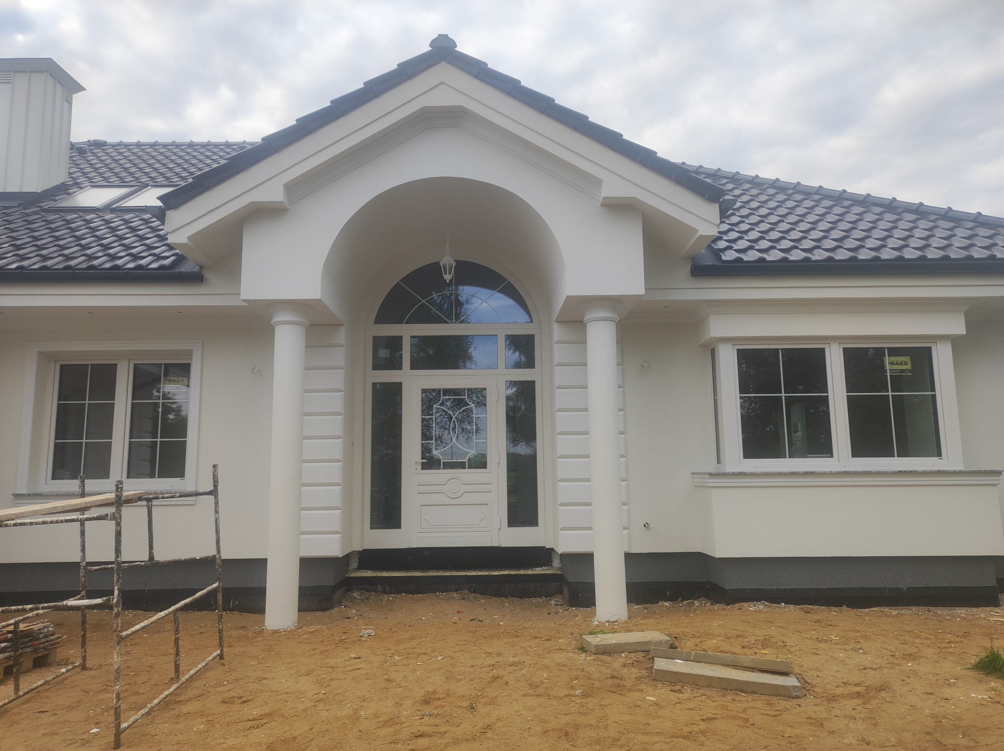 Biały dom jednorodzinny z kolumnami przy wejściu i ciemną dachówką, widok z przodu, teren wokół domu w trakcie prac wykończeniowych, pochmurne niebo.