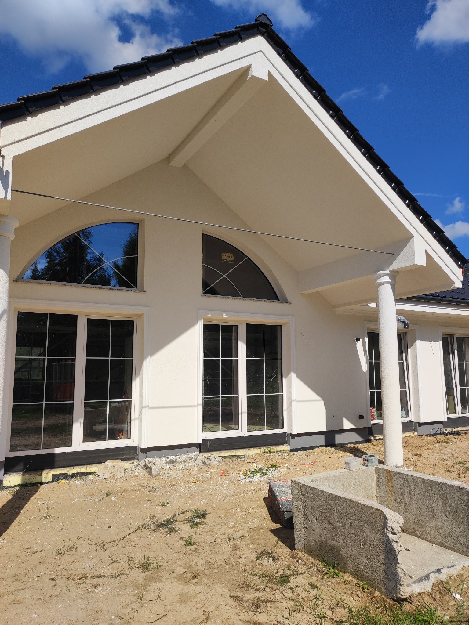 Elewacja domu jednorodzinnego z białym tynkiem, czarną dachówką i kolumnami. Widoczne okna z białymi ramami i elementami dekoracyjnymi. Dom w trakcie wykończenia.