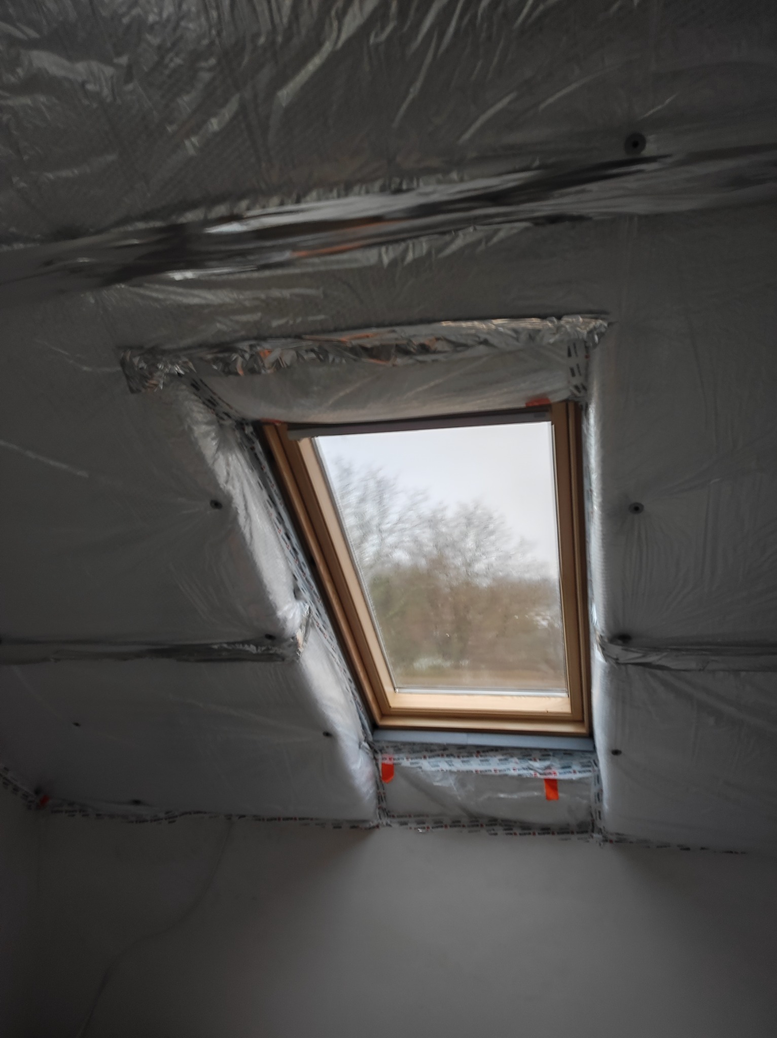 Poddasze z oknem dachowym w trakcie ocieplania, widoczna srebrna folia izolacyjna z taśmami uszczelniającymi na połączeniach, na zewnątrz widok na drzewa.
